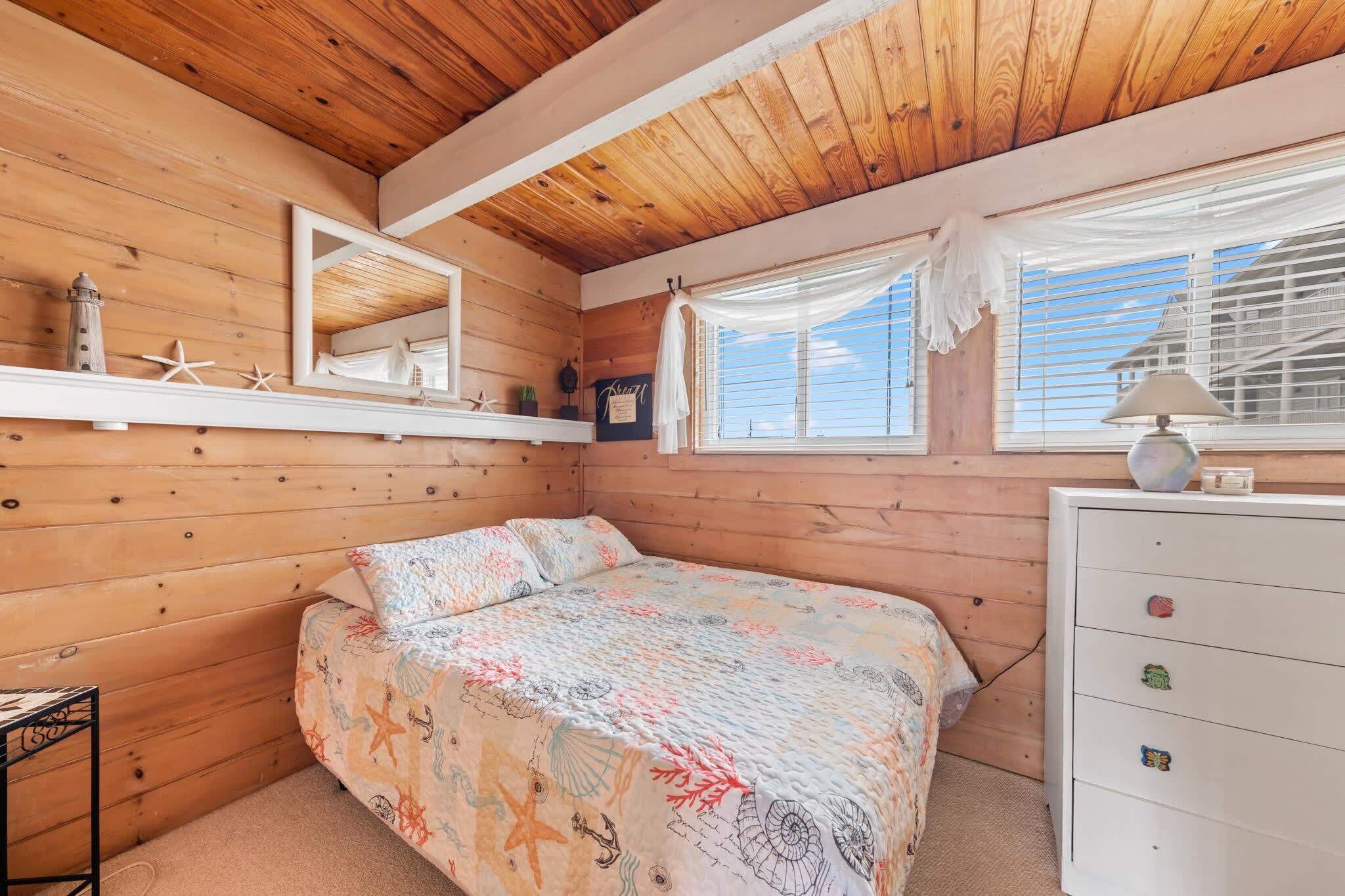 A cozy bedroom with a wooden ceiling, a bed with a colorful quilt, a dresser with drawers, a mirror, and a window with curtains.