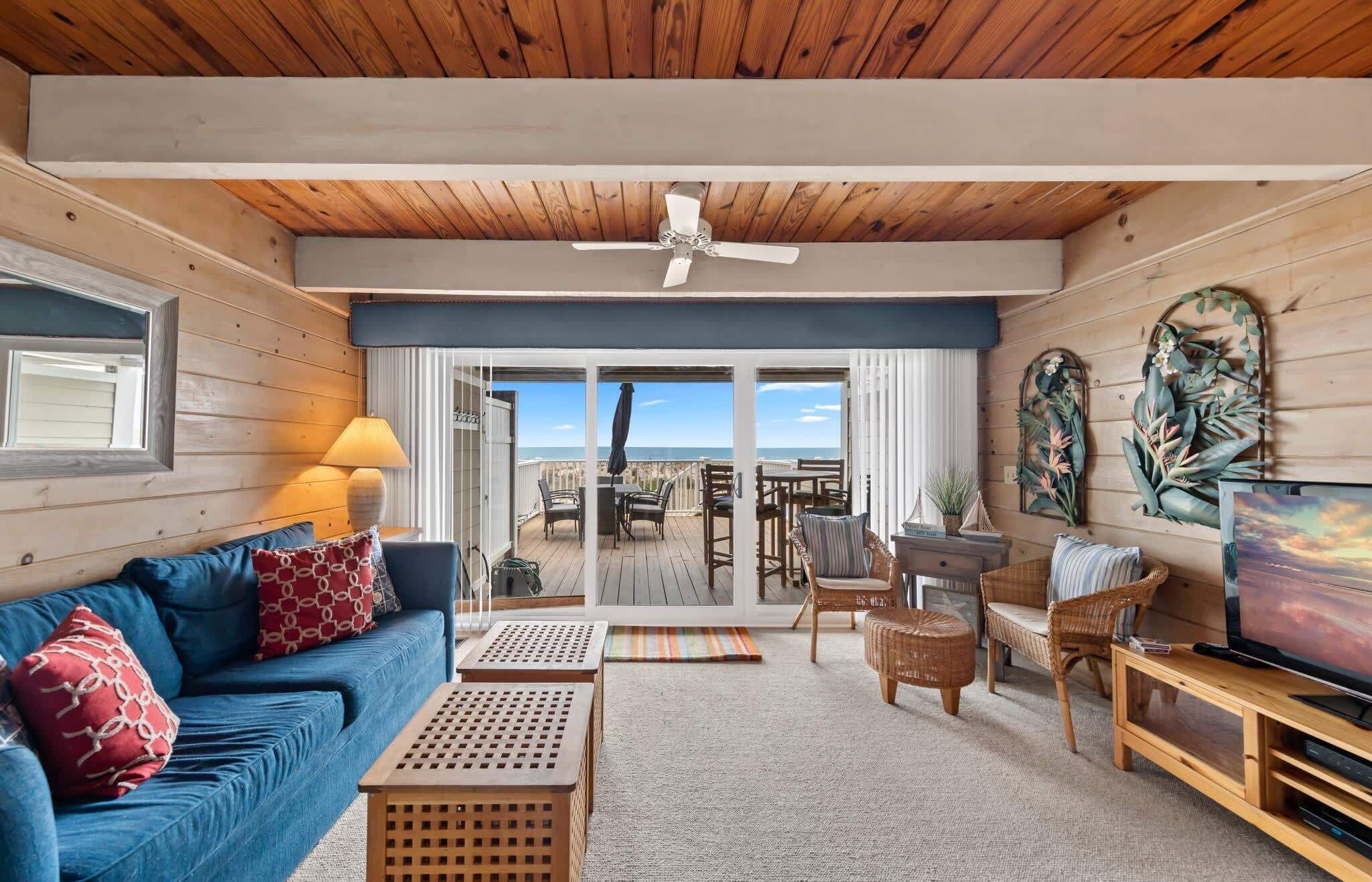 A cozy living room with a blue sofa, wooden coffee table, and a television. There is a balcony with a view of the ocean.