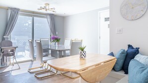 A modern living room with a wooden dining table, white chairs, and a sofa with blue cushions. A large clock is mounted on the wall. - Perfect for Multiple Families- Close to Beach, Trails, Wineries, Breweries (South Haven)