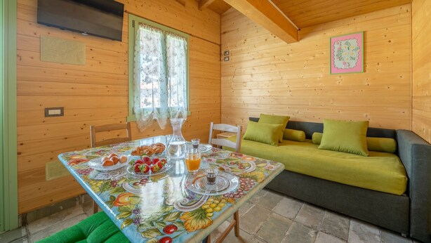 Interior de una cabaña de madera con una mesa de comedor lista para el desayuno, un sofá con cojines verdes y un televisor montado en la pared.