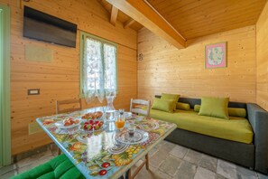 A wooden cabin interior with a dining table set for breakfast, a sofa with green cushions, and a television mounted on the wall.