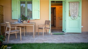 Un patio al aire libre con una mesa y sillas de madera, una puerta con contraventanas verdes y un pequeño jardín.