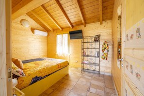 Ein Zimmer in einer Holzhütte mit einem Bett, einem Fernseher, einem Metallregal mit Büchern und einem bunten Schal sowie einem Holzschrank mit dekorativen Aufklebern.