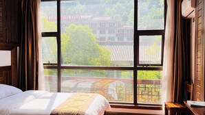 A hotel room with two beds, a large window with a view of traditional buildings and greenery, and a wooden ceiling. - zhuiyikezhan (Zhangjiajie)