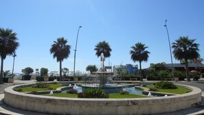 Une fontaine circulaire avec des jets d’eau, entourée de palmiers et surplombée d’un ciel bleu clair.