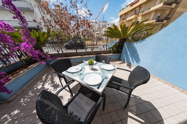 A patio with a table set for four, surrounded by wicker chairs, with a view of a building and a street in the background.