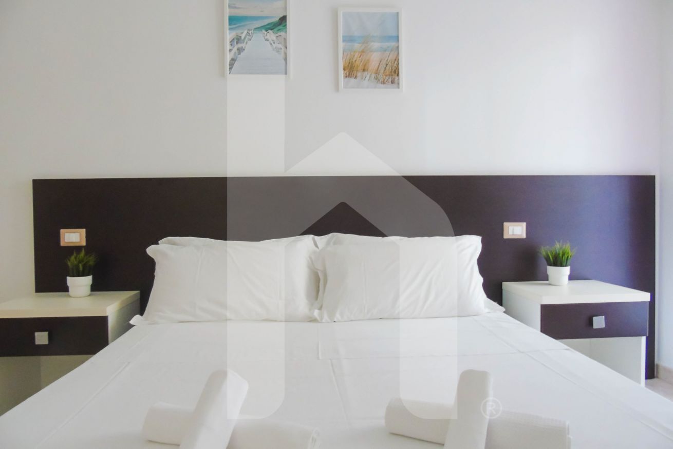Une chambre d’hôtel moderne avec un grand lit, du linge de lit blanc, deux tables de chevet ornées de plantes et des œuvres d’art accrochées aux murs.