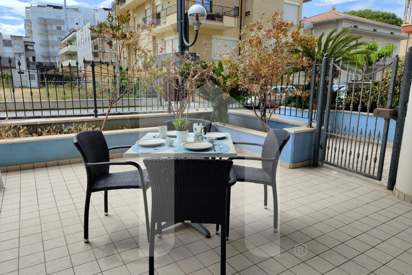 Une terrasse avec une table dressée pour quatre personnes, entourée de chaises en osier noir, et un petit arbre au centre.