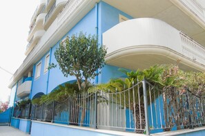 A multi-story building with balconies, a blue facade, and a metal fence with plants.
