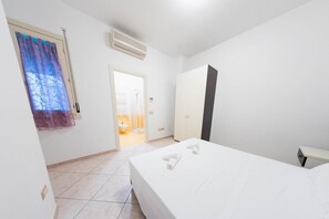 A hotel room with a bed, a wardrobe, a window with curtains, an air conditioning unit, and a door leading to a bathroom.