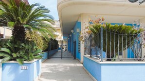 A narrow pathway flanked by blue walls and palm trees, leading to a building entrance with a metal gate. - Homiday - Felicioni Alba Adriatica, Bilocale al Piano Terra (Alba Adriatica)