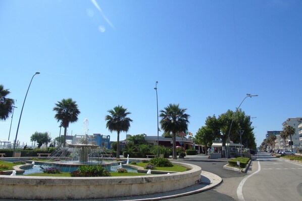A public square with a central fountain, palm trees, and a street with buildings in the background.