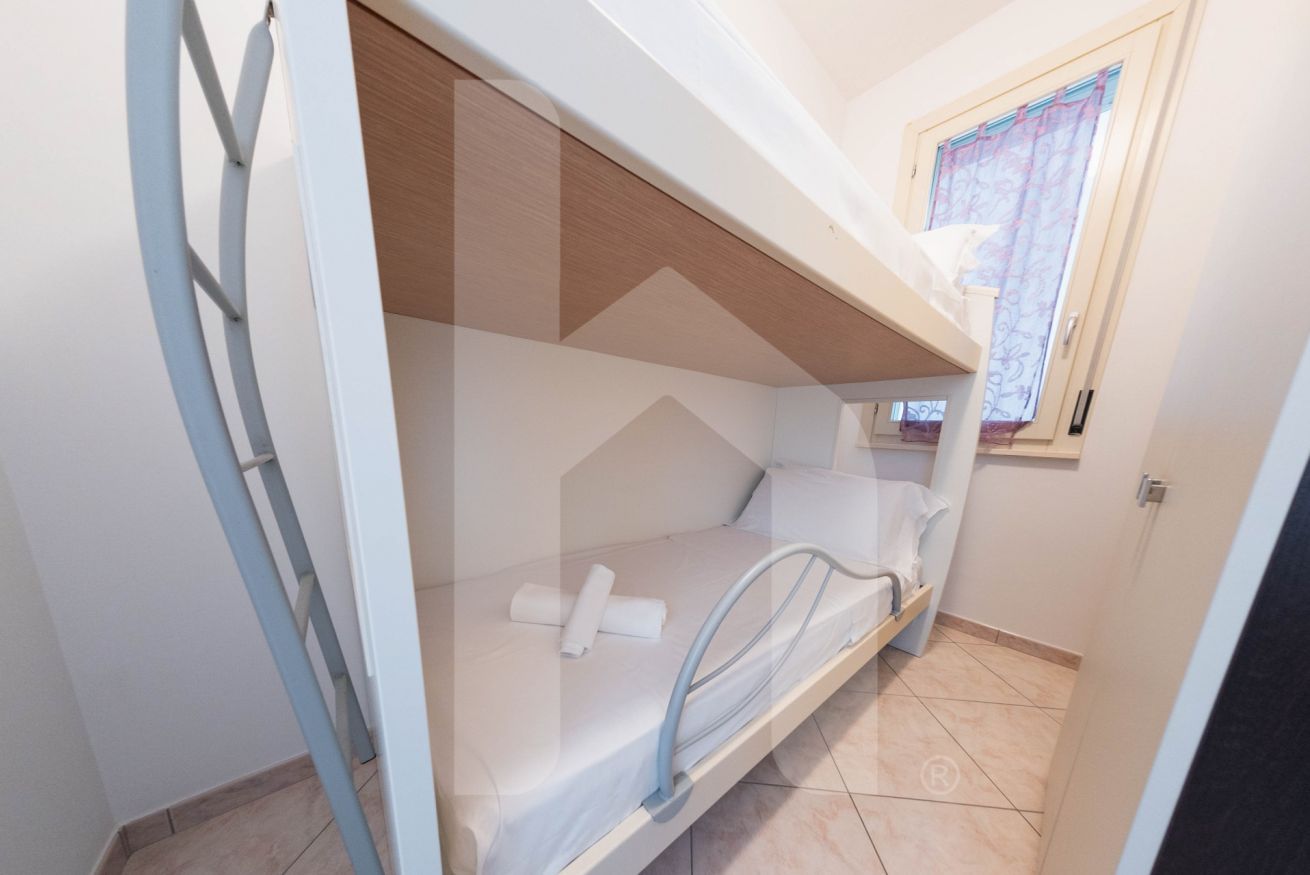 Habitació senzilla amb llit superposat, una finestra amb cortines i una porta.