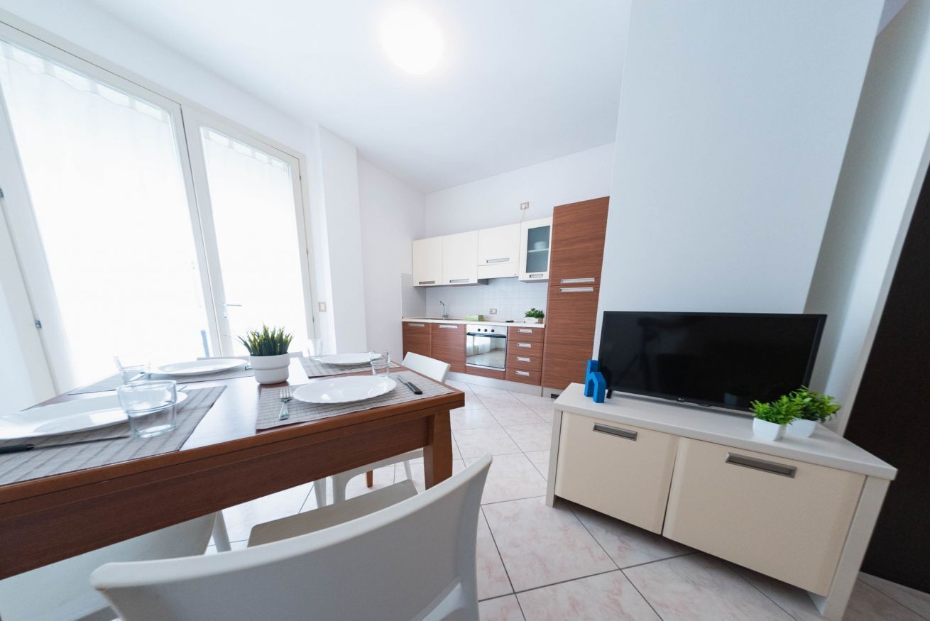 A modern kitchen with white cabinets, a dining area with a wooden table, and a TV on a cabinet.
