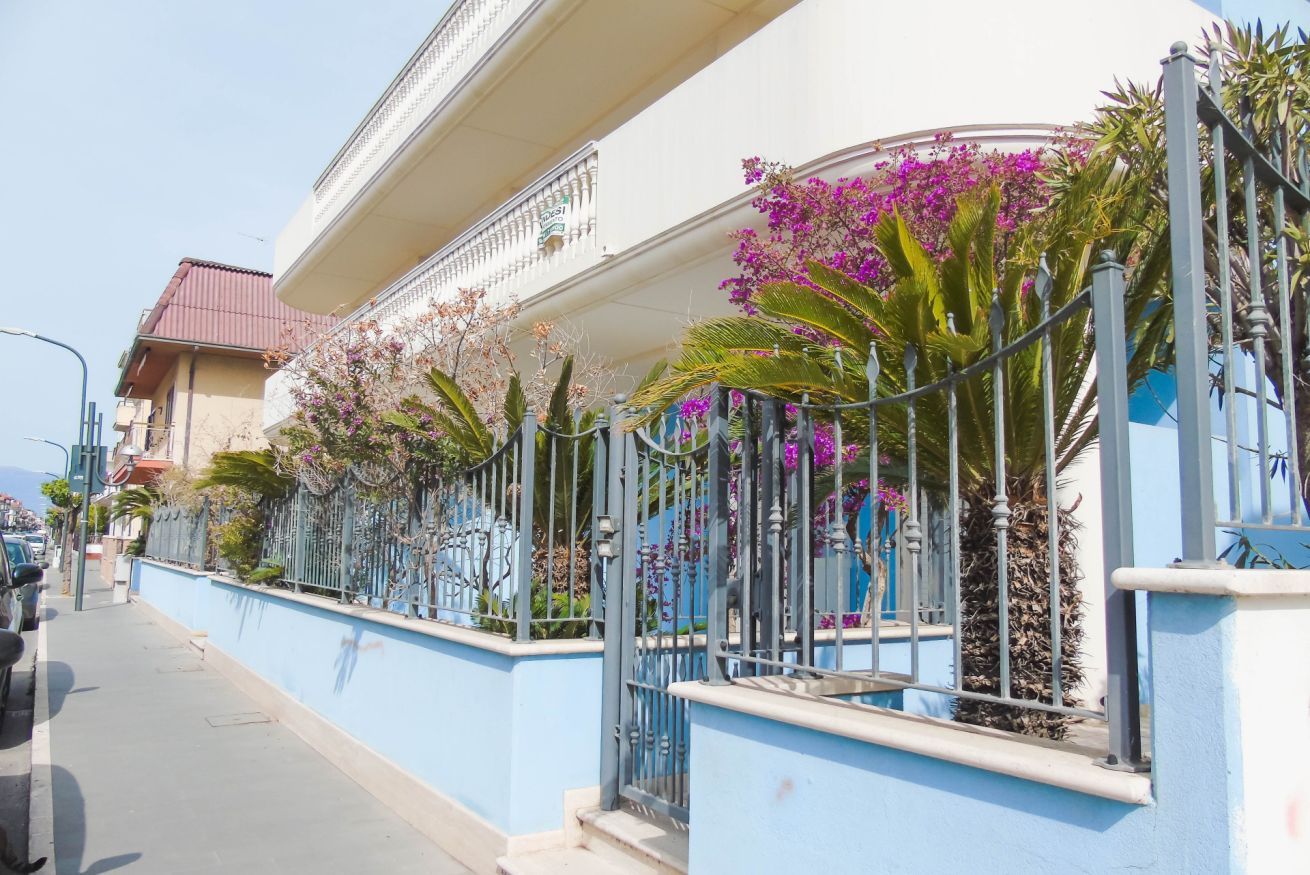 A residential building with a blue exterior, a balcony with purple flowers, and a metal fence.