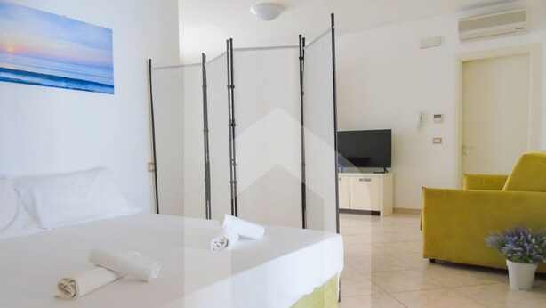 A hotel room with a bed, a TV, a yellow chair, and a painting of a sunset. - Homiday - Felicioni Alba Adriatica, Monolocale al 1°piano (Alba Adriatica)