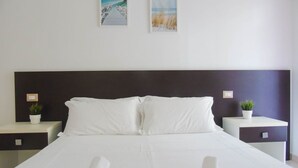 A neatly made bed with white linens, two bedside tables with potted plants, and two framed pictures on the wall.