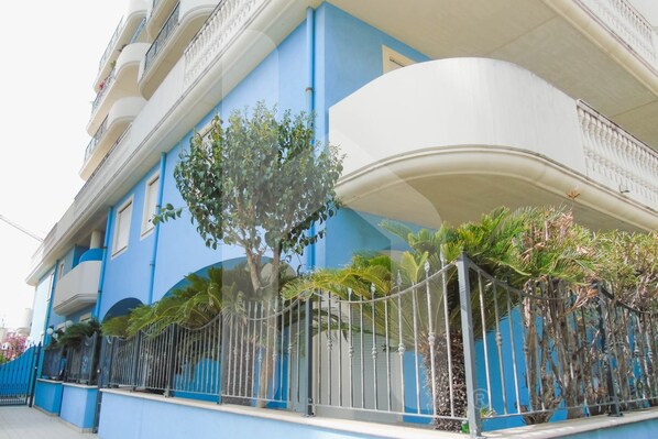 Un immeuble à plusieurs étages doté de balcons, une structure blanche aux lignes courbes et une clôture métallique ornée de plantes.
