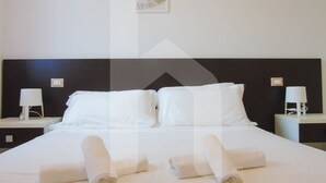 Un lit bien fait, avec du linge de lit et des serviettes blanches, deux tables de chevet avec des lampes et un tableau encadré au mur.