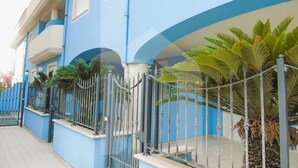 A row of blue buildings with balconies, a gated entrance, and palm trees.