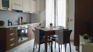 A modern kitchen with white cabinets, a built-in oven, a dining table with chairs, and a window with curtains.