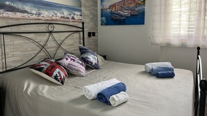 A neatly made bed with pillows and towels, a painting of a coastal scene, and a window with sheer curtains. - Brand new apartment near the sea (Fuscaldo)