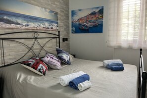 Un lit bien fait, avec des oreillers et des serviettes, un tableau représentant un paysage côtier et une fenêtre ornée de rideaux légers.