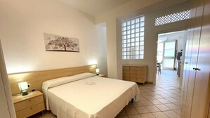 A bedroom with a bed, nightstand, wall art, window, and a door leading to another room. - Dante Apartment (Porto Azzurro)