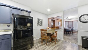A modern kitchen with a black refrigerator, wooden dining table, and chairs. There is a wall with a 'Live Love Laugh' sign and a clock. - Experience Wichita living near WSU!  Fire Pit, Pool Table, and Arcade games. (Wichita)