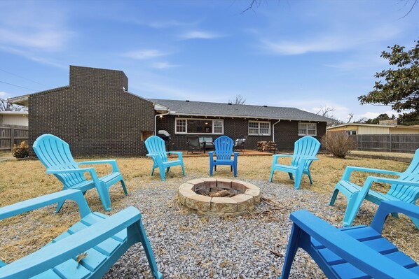 A backyard with a fire pit, four blue Adirondack chairs, and a brick house.