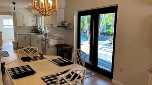 A dining area with a checkered table, white chairs, a chandelier, and a view of a pool through a glass door.