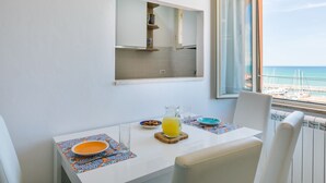 Zona pranzo con tavolo apparecchiato per due, vista sul mare attraverso una finestra e angolo cottura visibile nel riflesso dello specchio.