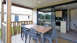 A covered patio with a wooden table and gray chairs, a sliding glass door leading to a kitchen area with a microwave and oven, and a view of a house in the background.