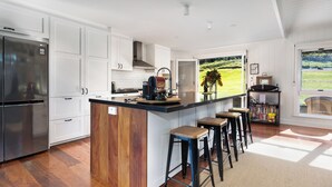 Una cocina moderna con armarios blancos, una isla de madera, taburetes de bar, una cafetera integrada y vistas a un paisaje verde a través de un ventanal grande.