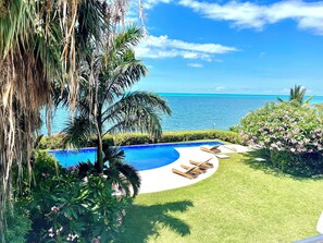 A pool with a curved edge, surrounded by well-maintained grass and palm trees, with a view of the ocean.