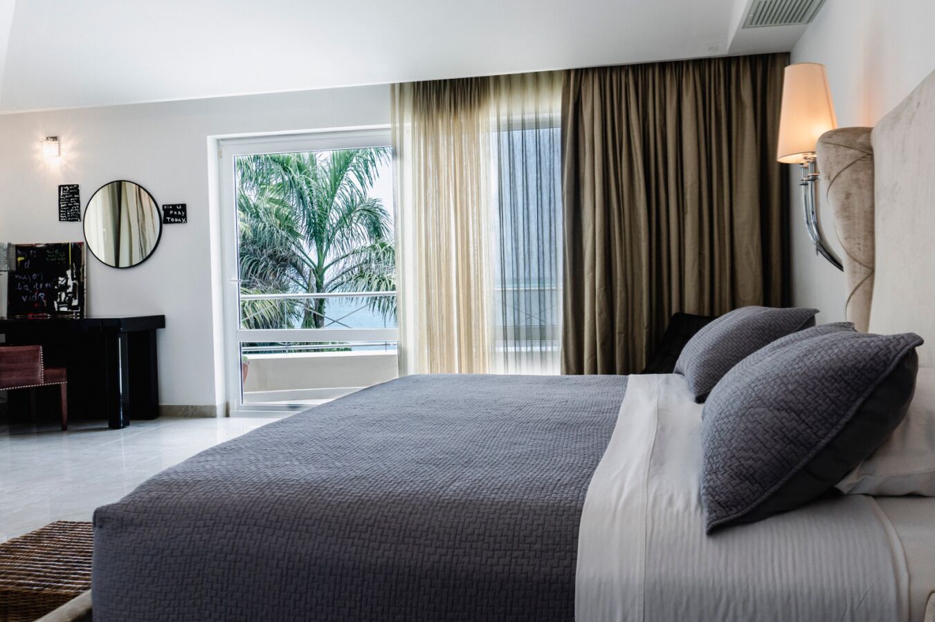 Ein Hotelzimmer mit einem großen Bett, einem Schreibtisch, einem Stuhl, einem Spiegel und Blick auf Palmen durch das Fenster.