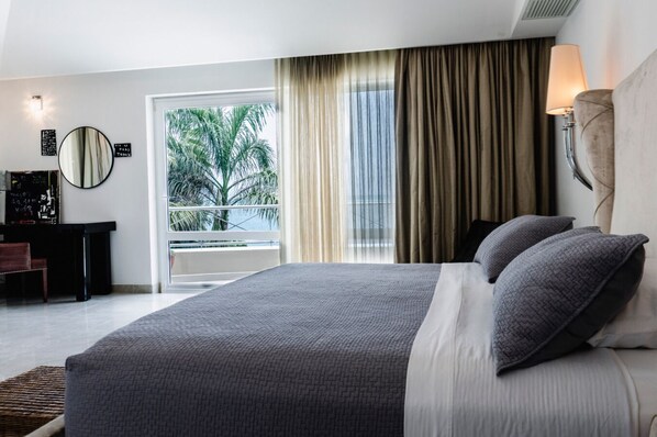 A hotel room with a large bed, a desk, a chair, a mirror, and a view of palm trees through the window.
