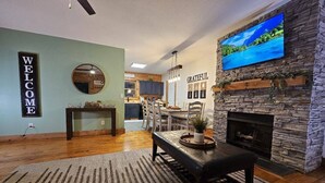 A modern living room with a stone fireplace, a television mounted on the wall, and a "WELCOME" sign. - Saluda Summit at Saluda Grade Cabins! (Saluda)