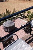 Un balcón con mesa y sillas, una zona de estar cubierta y vista a una zona arenosa con plantas.