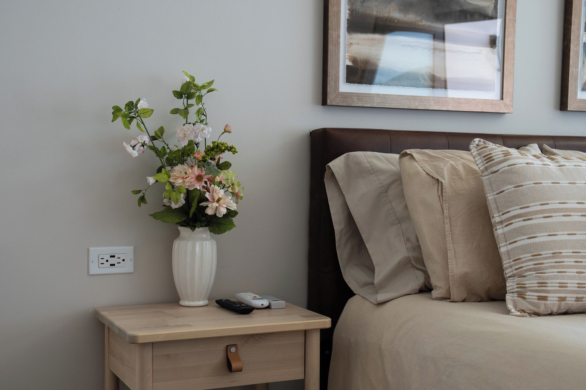 A neatly arranged bedroom with a bedside table, a vase of flowers, a bed with beige bedding, and a framed picture on the wall.