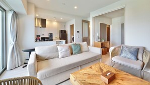 A modern living room with a sofa, a wooden coffee table, and a wicker chair. There is a kitchen area in the background with a microwave and a coffee machine.