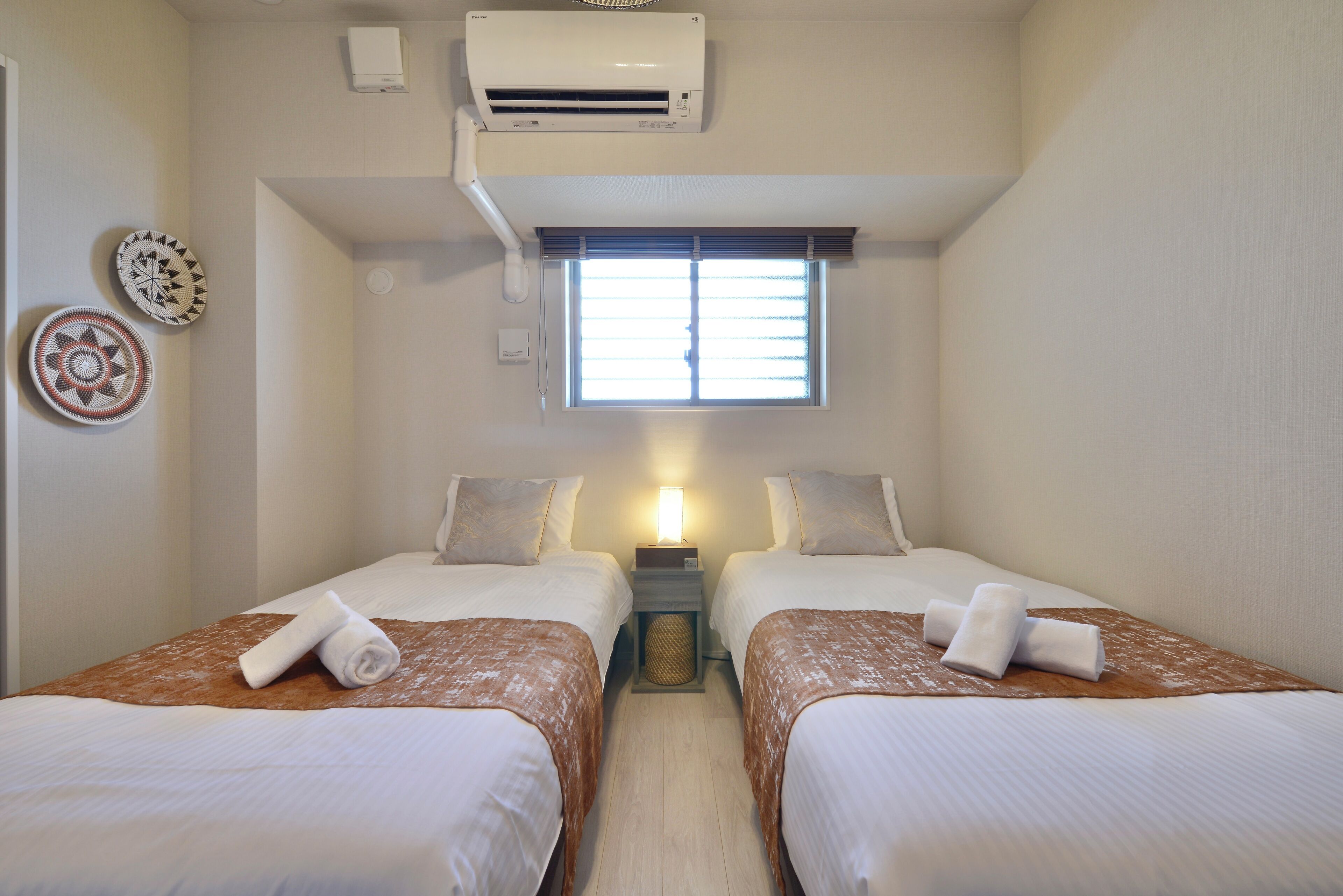 Ein Hotelzimmer mit zwei Betten, einem Fenster mit Jalousien, einer Klimaanlage und dekorativen Platten an der Wand.