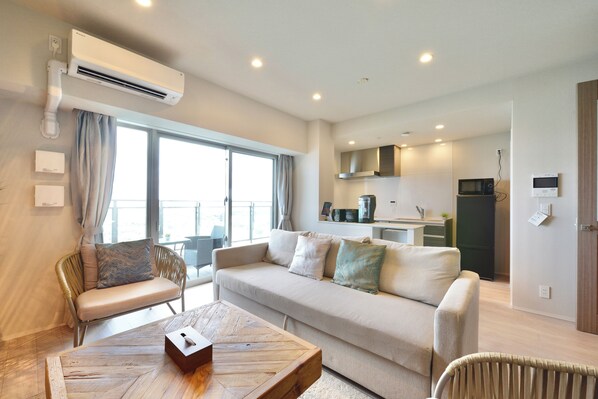 A modern living room with a sofa, a wooden coffee table, and a dining area with a kitchen in the background. - Branchera naha akebono premist R68 (Naha)