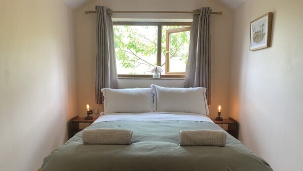 A neatly made bed with a green bedspread, two pillows, and two folded towels. There are two bedside tables with lamps, a window with curtains, and a framed picture on the wall.