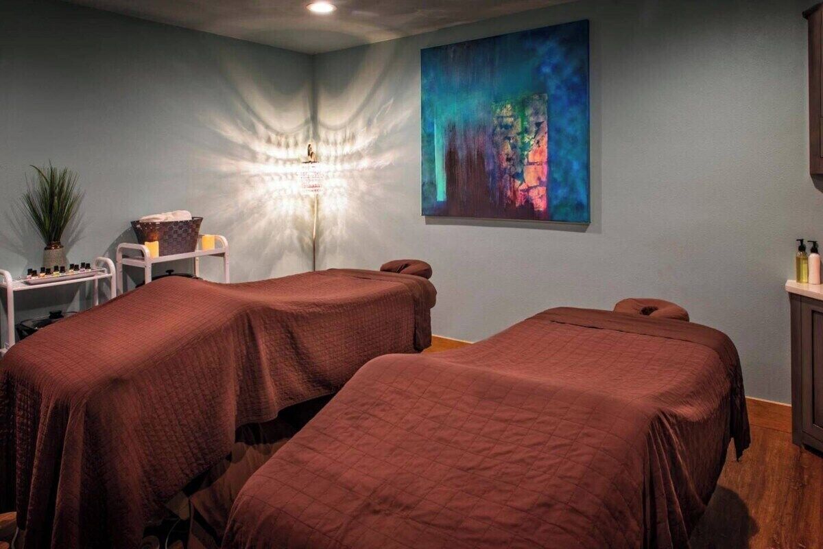 Ein Zimmer mit zwei Betten, einer Massageliege, einem an der Wand befestigten Massagesessel, einem kleinen Tisch mit Flaschen und einem großen abstrakten Gemälde.