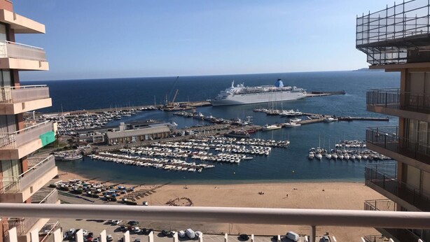 Ein Yachthafen mit zahlreichen Booten, ein großes Kreuzfahrtschiff und ein Strandabschnitt mit Personen und Liegestühlen.
