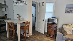Une cuisine avec une table et des chaises blanches, un four à micro-ondes posé sur un meuble, et une pancarte sur laquelle on peut lire : « CHAQUE ÉTÉ A SON HISTOIRE ».