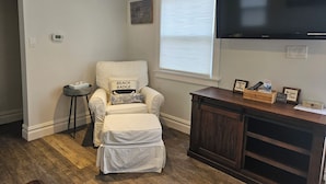 Un salon avec un fauteuil blanc, un petit pouf, une table d’appoint en bois agrémentée d’objets décoratifs, un téléviseur à écran plat et une fenêtre avec des stores.
