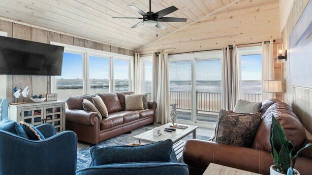 A room with a ceiling fan, a brown leather sofa, a blue armchair, a white coffee table, and a TV mounted on the wall. - Salty Kisses & Starfish Wishes by Brunswick VR: Oceanfront & Pet-Friendly (Ocean Isle Beach)