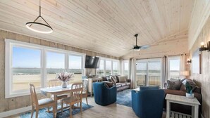 A spacious living area with a wooden ceiling, large windows, a dining table, and blue furniture. - Salty Kisses & Starfish Wishes by Brunswick VR: Oceanfront & Pet-Friendly (Ocean Isle Beach)
